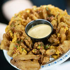 a fried onion and dip topped with bacon bits, cheese sauce and green onion from Colossal Onion 