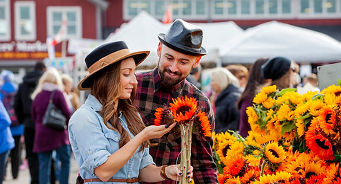 A couple admires orange sunflowers with a bustling crowd and St. Jacobs Farmers Market Building in the background 