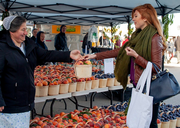 A woman with long red hair and a green scarf buys peaches from a vendor under an outdoor market tent. The vendor, wearing a bonnet and a dotted jacket, smiles as she hands the woman a basket of peaches. Tables filled with fruit are displayed in the background.